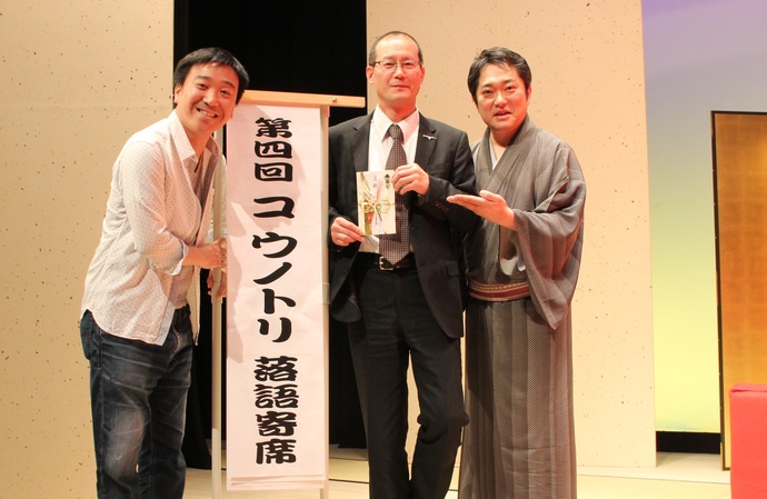 写真：舞台上に並ぶ林家菊丸師匠と月亭八光さんと寄付目録を手にした宮下課長