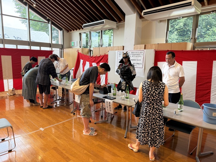 市民ホールに甲冑武者の衣装を着た武将ときき酒大会の選手が利き酒をしている。